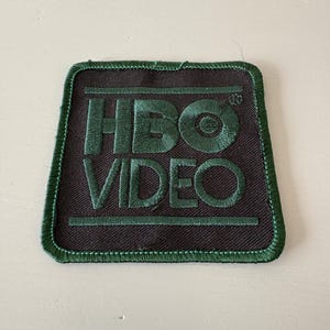May include: Embroidered patch featuring the text "HBO VIDEO" in dark green thread on a dark gray background. The patch has a green border and a square shape with slightly angled sides. The patch is likely for clothing or accessories.