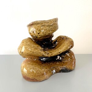 May include: A handcrafted ceramic sculpture with a stacked, organic form. The piece features a glossy, speckled glaze in shades of brown and tan, with darker accents. The sculpture has a unique, abstract design, suitable for home decor.