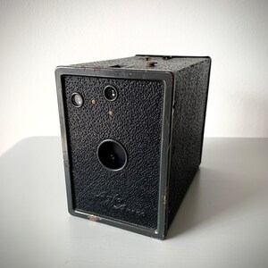 May include: A black vintage box camera with a textured finish. The camera has a round lens and two small viewing holes.