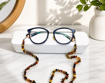 May include: Dark blue eyeglasses with thin metal temples resting on a white rectangular case. Attached to the frames is a long beaded chain made of small, polished, translucent amber-colored stones in varying shades of brown and golden yellow.