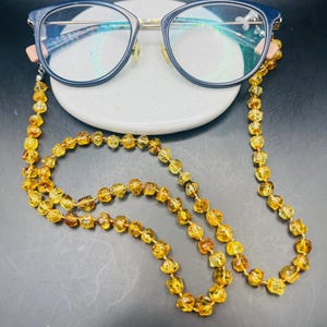 May include: Eyeglasses with a dark blue frame and a gold-colored bridge rest on a white ceramic dish. A necklace made of amber-colored beads is attached to the glasses. The beads are a mix of light and dark amber.