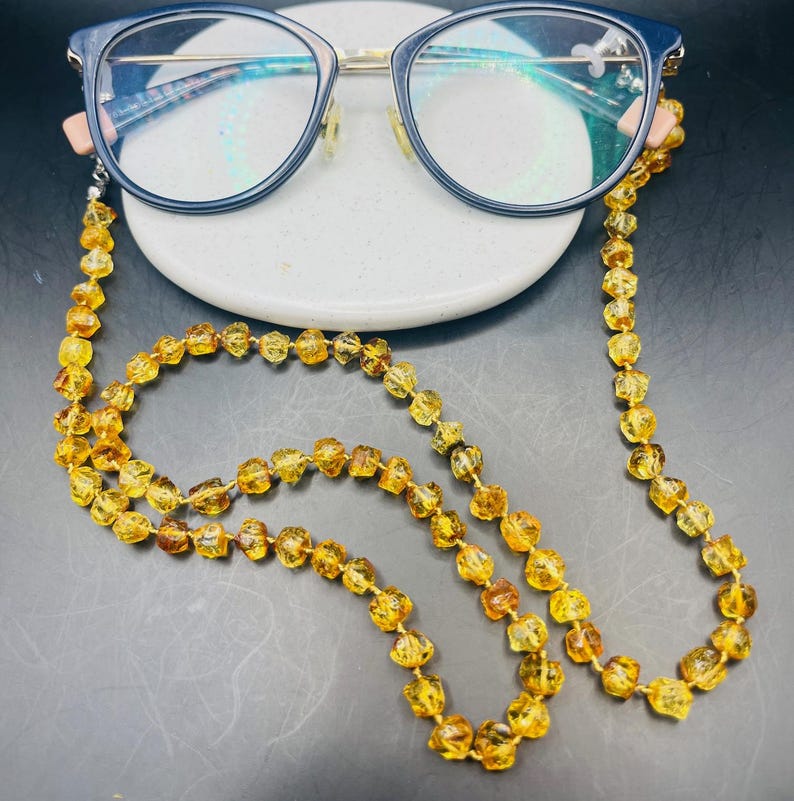 May include: Eyeglasses with a dark blue frame and a gold-colored bridge rest on a white ceramic dish. A necklace made of amber-colored beads is attached to the glasses. The beads are a mix of light and dark amber.