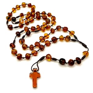 Puede incluir: Un collar con cuentas de color ámbar y un colgante de cruz. Las cuentas varían en tamaño y tono, desde amarillo claro hasta marrón oscuro. La cruz es de un color ámbar similar. El collar está ensartado en un cordón marrón oscuro.