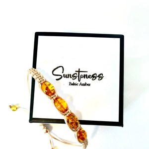 Pode incluir: Um bracelete de macram&eacute; bege com quatro contas de &acirc;mbar. O bracelete est&aacute; sobre um fundo branco com uma caixa preta que diz "Sunstoness Baltic Amber".
