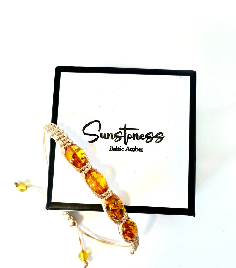 Pode incluir: Um bracelete de macram&eacute; bege com quatro contas de &acirc;mbar. O bracelete est&aacute; sobre um fundo branco com uma caixa preta que diz "Sunstoness Baltic Amber".