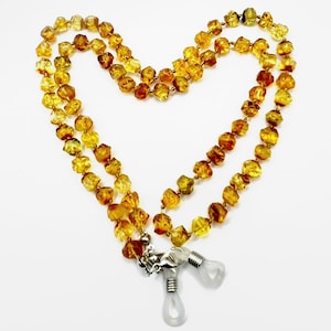 May include: A necklace made of amber-colored, irregularly shaped beads. The necklace is designed to hold eyeglasses, with a silver-colored clasp and clear rubber loops. The beads are a mix of golden and brown hues, creating a warm, natural aesthetic.