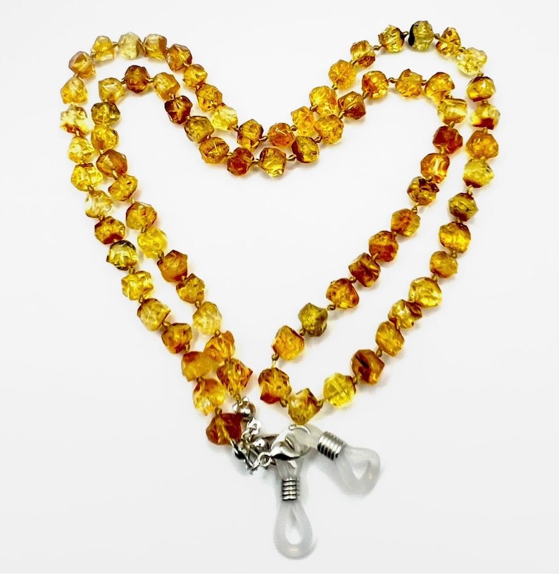 May include: A necklace made of amber-colored, irregularly shaped beads. The necklace is designed to hold eyeglasses, with a silver-colored clasp and clear rubber loops. The beads are a mix of golden and brown hues, creating a warm, natural aesthetic.