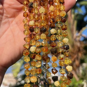 May include: A multi-strand eyeglass chain featuring irregularly shaped amber beads in shades of yellow, brown, and cream. The chain has silver-colored clasps and loops for attaching to glasses. The background shows green foliage and blue sky.
