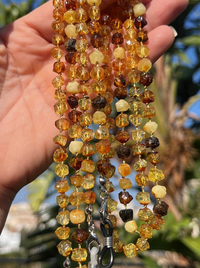 May include: A multi-strand eyeglass chain featuring irregularly shaped amber beads in shades of yellow, brown, and cream. The chain has silver-colored clasps and loops for attaching to glasses. The background shows green foliage and blue sky.