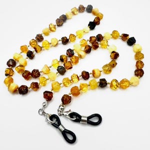 May include: A multi-colored amber beaded eyeglass chain. The beads are various shades of brown, yellow, and cream. The chain has silver-colored clasps and black rubber loops to attach to glasses. The chain is laid out on a white surface.