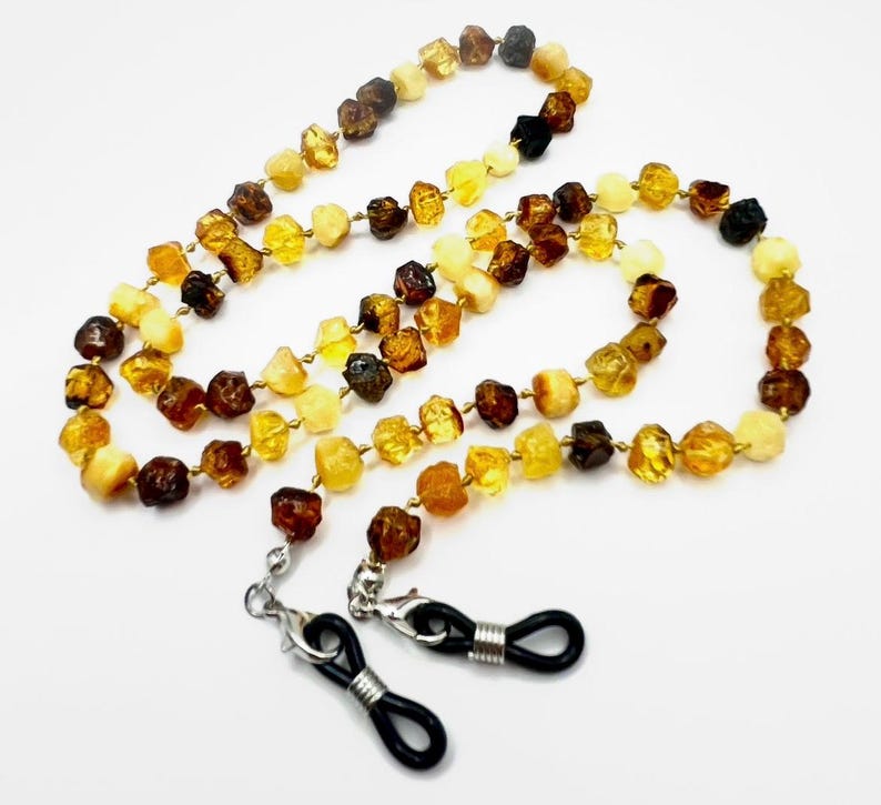 May include: A multi-colored amber beaded eyeglass chain. The beads are various shades of brown, yellow, and cream. The chain has silver-colored clasps and black rubber loops to attach to glasses. The chain is laid out on a white surface.