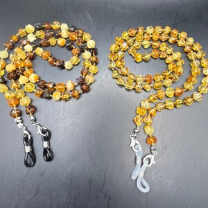May include: Two beaded eyeglass chains. One chain features a mix of amber and brown beads, while the other has amber-colored beads. Both chains have silver-colored clasps and black or clear rubber loops for attaching to glasses.