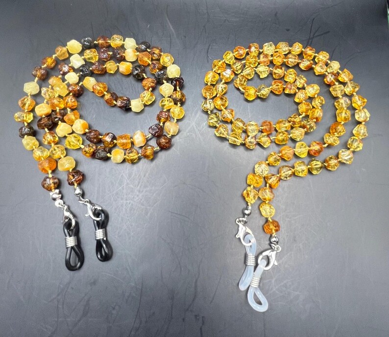 May include: Two beaded eyeglass chains. One chain features a mix of amber and brown beads, while the other has amber-colored beads. Both chains have silver-colored clasps and black or clear rubber loops for attaching to glasses.