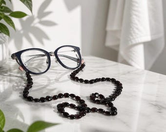 May include: A pair of dark blue eyeglasses with a long, beaded chain attached to the temples. The chain consists of numerous small, dark, oval-shaped beads strung together in a continuous line, resting in a curved pattern below the frames.