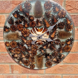 May include: A round clock with a blue and white resin background. The clock face is decorated with pine cones and other natural elements. The clock hands are black metal.