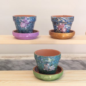 May include: Three small terracotta flower pots with colourful, abstract designs and gold fish accents. Each pot has a matching saucer in a different colour: purple, gold, and green.
