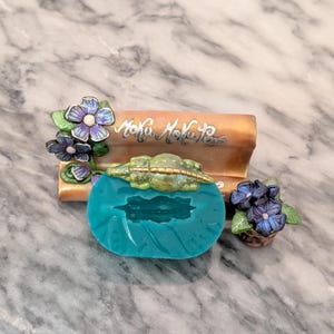 May include: A teal silicone mould with a small alligator design. The mould is sitting on a brown and gold base with the words "Moku Moku Press" written on it. There are also two small flower arrangements with purple and blue flowers.