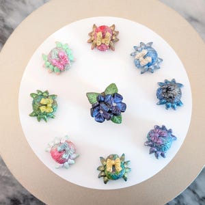 May include: Nine handmade polymer clay flower brooches in a variety of colours and designs. The brooches feature a range of flowers, including hydrangeas, roses, and daisies. Each brooch has a unique colour scheme and is adorned with a small bow.