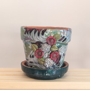 May include: A decorative terracotta flower pot with a green and white marbled design. The pot has a saucer and is decorated with red and silver flowers.