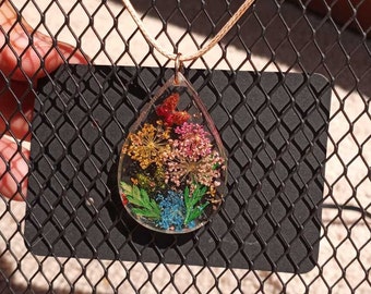 Puede incluir: Un collar con un colgante en forma de lágrima, con un marco de resina transparente que contiene un surtido colorido de flores prensadas y purpurina.