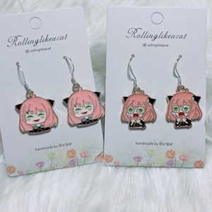 May include: Two pairs of dangle earrings featuring a cartoon character with pink hair, black cat ears, and a black and white outfit. The earrings are silver with a gold accent. The text "Rollinglikeacat" and "@rollinglikeacat" are on the card. The text "handmade by @ra'ra" is on the card.