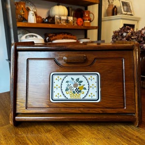 May include: A rectangular wooden bread box with a roll-top door and a central handle. The front panel features a decorative rectangular tile inlay displaying a colorful illustration of a fruit basket with yellow, green, and brown tones.