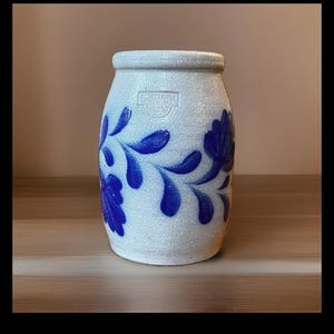 May include: A cylindrical stoneware crock with a textured off-white finish and a blue hand-painted floral vine pattern. The vessel features a stamped maker's mark near the rim and a slightly tapered shape with a smooth, rounded top edge.