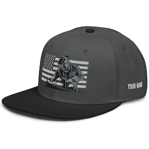 Può includere: Un cappello da baseball grigio scuro e nero con un'immagine ricamata di un saldatore che lavora davanti a una bandiera americana. Il testo "YOUR NAME" è ricamato sul retro del cappello.