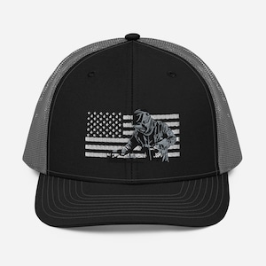 Puede incluir: Una gorra de camionero negra y gris con una bandera estadounidense bordada en blanco y un soldador trabajando en metal. La bandera está detrás del soldador.