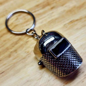 May include: A silver-colored keychain featuring a miniature welding helmet. The helmet has a rectangular black visor and a textured, grid-like pattern on the lower portion. The keychain includes a silver ring for attaching keys.