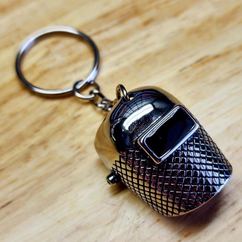 May include: A silver-colored keychain featuring a miniature welding helmet. The helmet has a rectangular black visor and a textured, grid-like pattern on the lower portion. The keychain includes a silver ring for attaching keys.