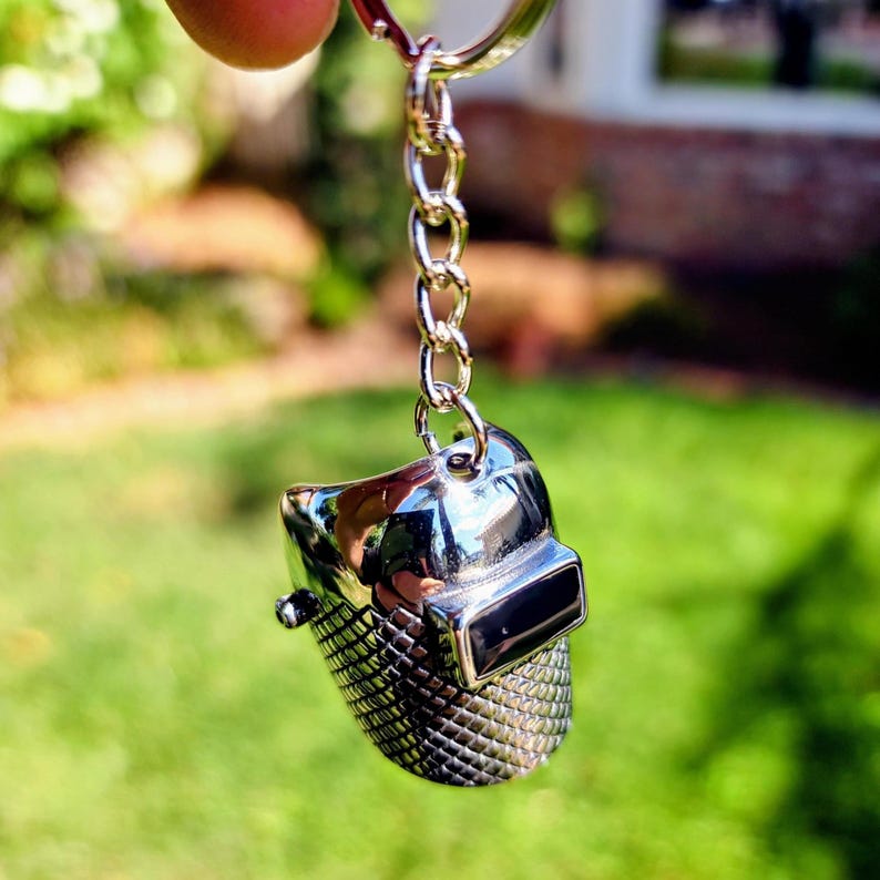 May include: A silver-colored keychain featuring a miniature replica of a welding helmet. The helmet has a shiny, reflective surface and is attached to a silver chain and keyring. The keychain is held up against a blurred green background.