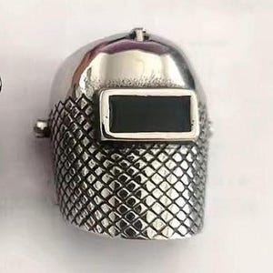 May include: A silver-colored ring shaped like a welding helmet. The ring has a shiny, smooth dome and a textured, diamond-patterned lower section. The rectangular visor is black. The ring is a unique piece of jewelry.