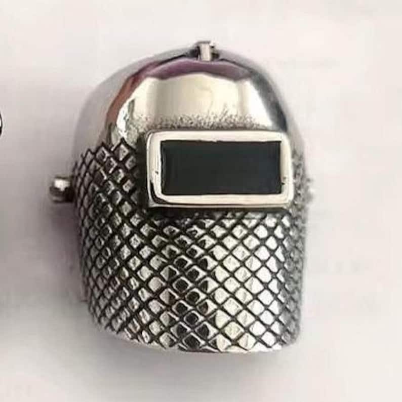 May include: A silver-colored ring shaped like a welding helmet. The ring has a shiny, smooth dome and a textured, diamond-patterned lower section. The rectangular visor is black. The ring is a unique piece of jewelry.