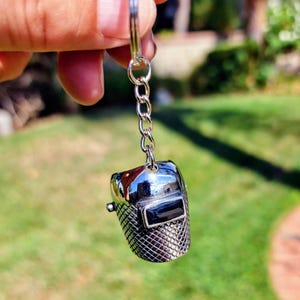 May include: A silver-colored keychain featuring a miniature welding helmet. The helmet has a shiny, reflective surface with a black rectangular visor. The lower portion of the helmet has a textured, grid-like pattern. The keychain is attached to a silver chain.
