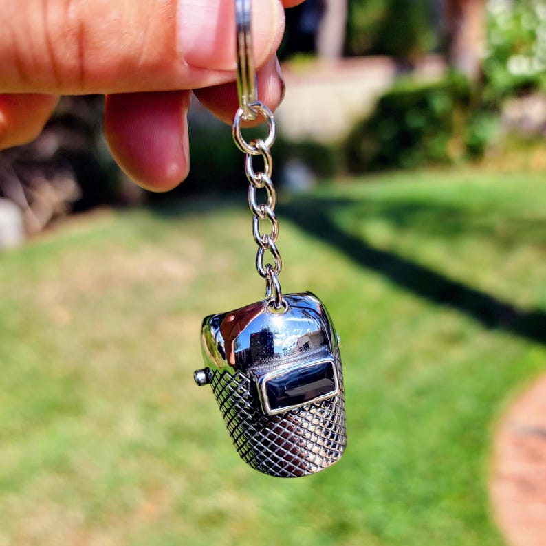 May include: A silver-colored keychain featuring a miniature welding helmet. The helmet has a shiny, reflective surface with a black rectangular visor. The lower portion of the helmet has a textured, grid-like pattern. The keychain is attached to a silver chain.