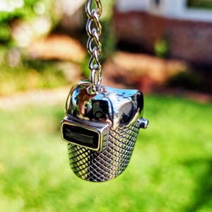 May include: A shiny silver-colored pendant in the shape of a welding helmet, hanging from a chain. The helmet has a detailed, textured lower section and a rectangular visor. The background is blurred green.