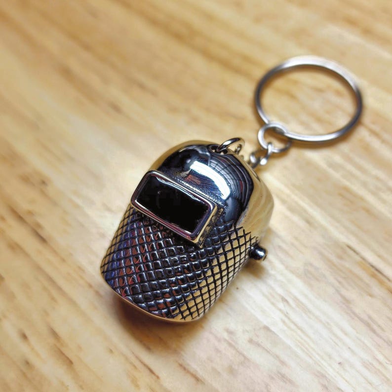 May include: A shiny silver-colored keychain featuring a miniature welding helmet. The helmet has a rectangular black visor and a textured, grid-like pattern on the lower portion. The keychain includes a metal ring for attaching keys.