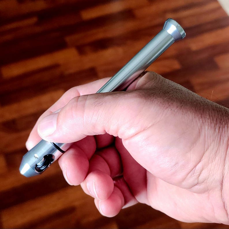 May include: A silver metal pen held in a hand. The pen has a cylindrical body with a slightly wider tip and a flared end. The pen appears to be made of a shiny, metallic material.