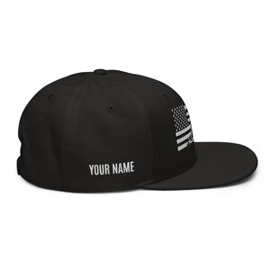 May include: Black and white American flag embroidered on a black baseball cap with a black brim. The cap has a white text label that says "YOUR NAME".