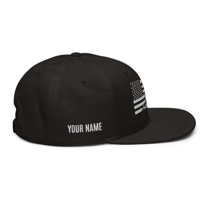 May include: Black and white American flag embroidered on a black baseball cap with a black brim. The cap has a white text label that says "YOUR NAME".