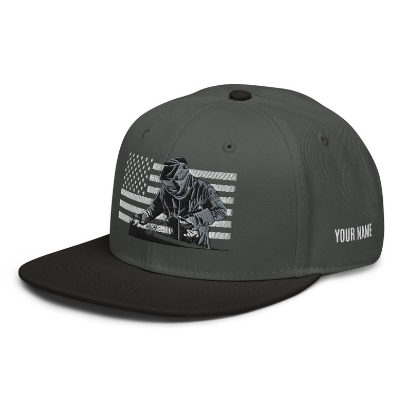 May include: A dark gray and black baseball cap with an embroidered image of a welder working in front of an American flag. The text "YOUR NAME" is embroidered on the back of the cap.
