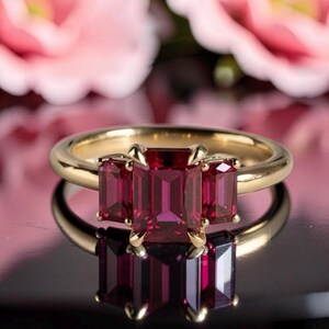 May include: A gold ring featuring three emerald-cut deep red gemstones set in a prong setting. The center stone is larger than the two flanking side stones, all mounted on a polished gold band that reflects on the dark surface below.