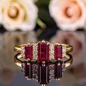 May include: A gold ring featuring a central rectangular baguette-cut red ruby flanked by two smaller rectangular red rubies, with small round white diamonds set between the stones in a symmetrical arrangement on a polished gold band.
