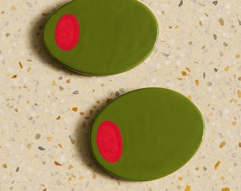 May include: Two olive-shaped coasters with a bright green base and a red dot, resembling olives. The coasters are placed on a speckled, light-colored surface, creating a playful and unique design.