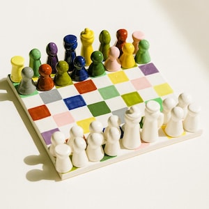 Puede incluir: Un juego de ajedrez de cerámica colorido con un tablero a cuadros. Las piezas de ajedrez son de varios colores, incluyendo amarillo, verde, azul y rojo. Las piezas blancas están en un lado, y las piezas coloridas en el otro.