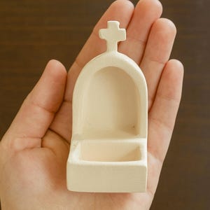 以下が含まれることがあります： 小さなオフホワイトの宗教的な壁掛け聖水盤。 聖水盤は上部に十字架があり、下部に小さな洗面器があります。 このアイテムは人の手で持たれています。