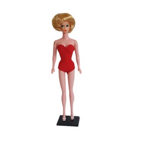 May include: A blonde doll wearing a red strapless swimsuit and standing on a black stand.