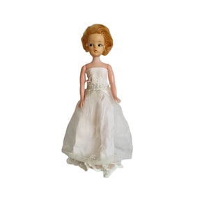 Könnte beinhalten: Eine vintage blonde Puppe in einem weißen Spitzen-Hochzeitskleid. Die Puppe hat ein bemaltes Gesicht und blaue Augen.