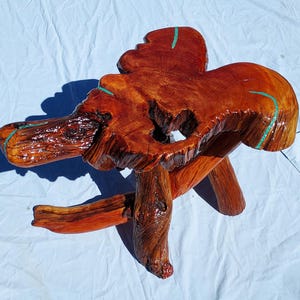May include: A handcrafted wooden table with a unique, organic shape. The table is made from a single piece of wood, with a rich, reddish-brown color and a glossy finish. Turquoise accents are inlaid. The table has three legs and a natural, rustic aesthetic.